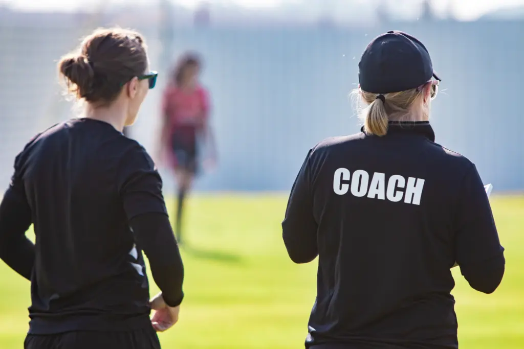 Rear view of two coaches watching players on a field.
