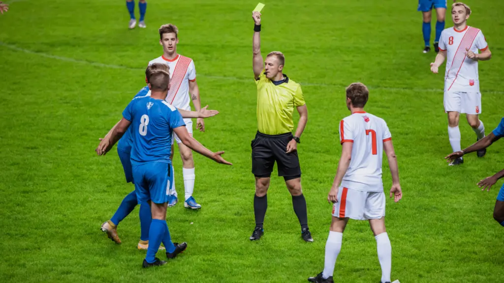 A referee holding up a yellow card and blowing a whistle during a soccer match.