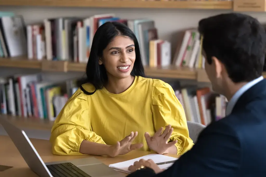A young woman in a yellow shirt talks to a man in a suit at a table in a school library.