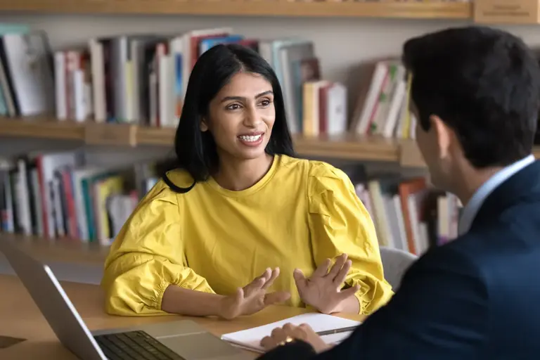 A young woman in a yellow shirt talks to a man in a suit at a table in a school library.